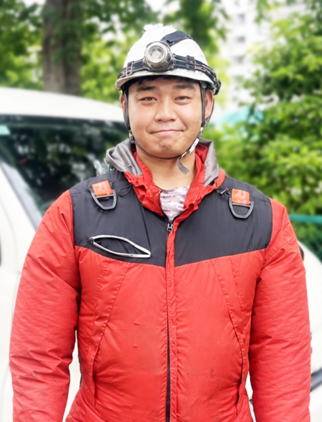 G.D.さんインタビューキービジュアル　屋外の作業車両前で、ライト付きヘルメットを装着し微笑むポートレート